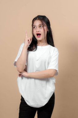 Uzun saçlı ve beyaz tişörtlü heyecanlı genç Asyalı kadın işaret parmağıyla işaret ederken şaşkınlık içinde, bej arka planda yeni bir fikir veya keşfi simgeliyor..