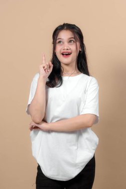 Uzun siyah saçlı heyecanlı bir Asyalı kadın beyaz tişört giyiyor, işaret parmağıyla gülümsüyor, harika bir fikir olduğunu söylüyor, bej arka planda duruyor..