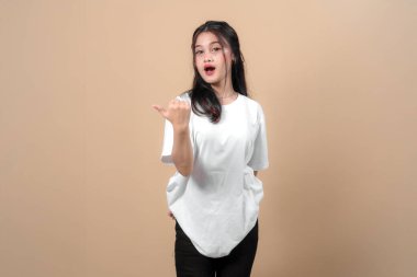 Beyaz tişört giyen genç Asyalı bir kadın, başparmağıyla kenarı işaret ederken şaşkınlığını dile getiriyor. Stüdyoda bej arka planda duruyor ve reklam için fotokopi alanı var..