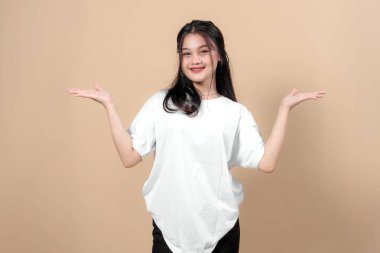 Beyaz tişörtlü genç Asyalı bir kadın gülümsüyor ve bej bir arka plana karşı elleri açık poz veriyor, neşeli bir ifadeyle bir şey sunuyor veya karşılaştırıyor..