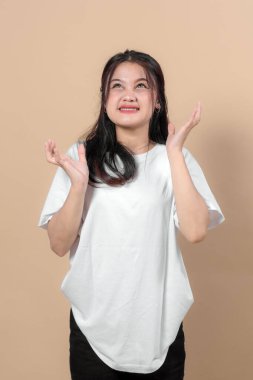 Beyaz tişörtlü genç Asyalı kadın umut dolu bir ifadeyle yukarı bakıyor. Sorgulamada ya da minnet duyarak ellerini kaldırıyor. Bej arkaplanlı stüdyo portresi.