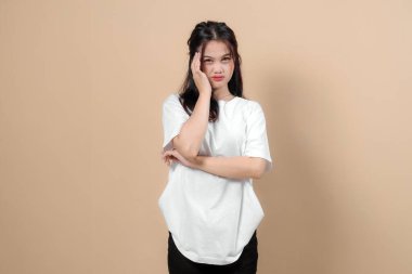 Beyaz tişörtlü genç Asyalı bir kadın acı dolu bir ifadeyle kafasına dokunuyor, muhtemelen baş ağrısı ya da stres yaşıyor. Sade bej arkaplanlı stüdyo çekimi.