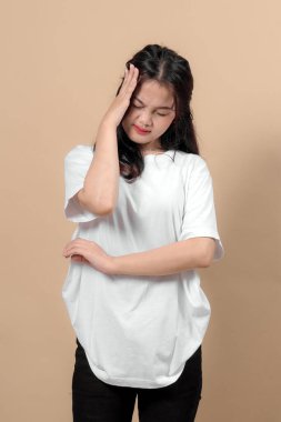 Beyaz tişörtlü, siyah kotlu, başını kapalı tutan, baş ağrısı ve stresi ifade eden, bej arka planda izole edilmiş, Asyalı genç bir kadın..