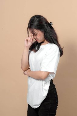 Beyaz tişörtlü ve siyah kotlu üzgün genç Asyalı kadın yan yana duruyor, yüzünü elle kapatıyor, stres ya da hayal kırıklığını ifade ediyor. Bej arka planda stüdyo portresi.