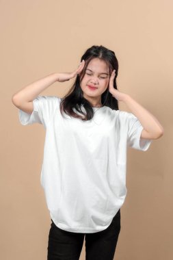 Beyaz tişörtlü Asyalı bir kadın bej arka planda şiddetli baş ağrısı, stres ya da hüsran belirtileri gösteriyor..