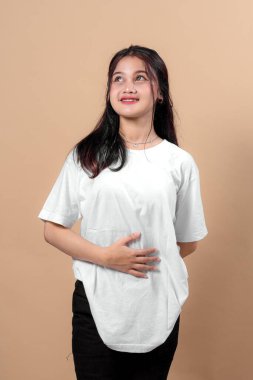 Beyaz tişörtlü genç bir Asyalı kadının portresi. Nazik bir gülümseme ve bir eli karnında bej arka planda umutlarını, minnettarlıklarını ya da memnuniyetlerini ifade ediyor..