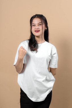 Beyaz tişörtlü mutlu genç Asyalı kadın heyecanla gülümsüyor, özgüvenini, coşkusunu, motivasyonunu ve zaferini stüdyo arka planında ifade ediyor..