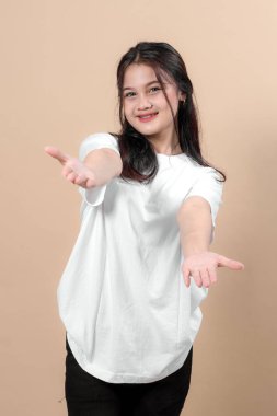 Beyaz tişörtlü neşeli genç Asyalı kadın gülümsüyor ve her iki koluna da hoş geldin pozu veriyor. Stüdyo ışığı altında tarafsız bir bej arka planda güvenle duruyor..