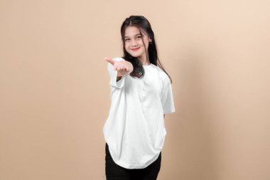 Beyaz tişörtlü, elini kameraya doğru uzatan Asyalı genç bir kadının portresi. Dostane bir gülümseme, davetkar bir jest ve bej arka planda sıcak bir ifade..