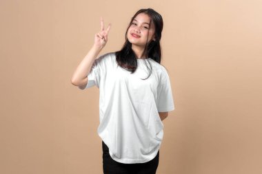 Düz beyaz tişört giyen genç Asyalı kadın barış işareti işareti gösterirken özgüvenle gülümsüyor, bej arka planda duruyor, sıradan bir poz veriyor..