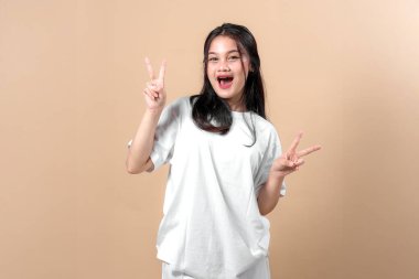 Heyecanlı genç Asyalı kadın, parlak bir gülümsemeyle iki elini kaldırarak çift barış işareti gösteriyor, beyaz büyük beden bir tişört giyiyor stüdyo ışıklandırmasında düz bir bej arka plan.