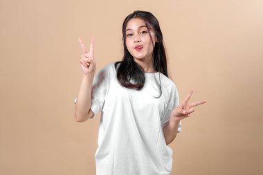 Beyaz tişörtlü oyuncu genç Asyalı kadın iki eliyle çifte barış işareti gösteriyor ve dilini uzatıyor. Stüdyoda bej arka planda sade bir şekilde poz veriyor..