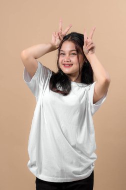 Neşeli, genç Asyalı kadın başının üstünde çifte barış işareti jesti ile poz veriyor, düz beyaz tişört ve siyah pantolon giyiyor, bej arka planda gülümsüyor..