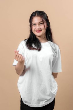 Mutlu genç Asyalı bir kadın Kore 'li bir parmak kalp hareketi yapıyor. Tek eliyle. Sade beyaz bir tişört giyerken içten bir şekilde gülümsüyor..