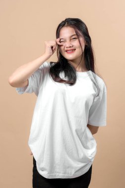 Beyaz büyük tişörtlü neşeli bir Asyalı kadının portresi, gülümseyen ve göz kırparken sevimli bir parmak kalp hareketi yapıyor, bej bir arka plana karşı..