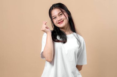 Sade beyaz tişört giyen gülümseyen Asyalı genç bir kadın bej arka planda duruyor. Koreli bir parmak kalp hareketi gösteriyor. Neşeli ve dostane bir yüz ifadesi var..