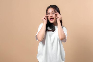 Büyük beyaz tişörtlü heyecanlı bir Asyalı kadın iki eliyle çift parmaklı kalp hareketi yapıyor, ağzı açık gülümsüyor, sağlam bir bej arka planda duruyor..