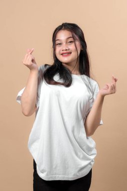 Beyaz tişörtlü mutlu bir Asyalı kadın gülümsüyor ve iki eliyle kalp hareketi yapıyor, bej bir arka plan önünde şakacı bir şekilde poz veriyor, neşeli görünüyor..