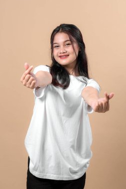 Beyaz tişörtlü neşeli genç Asyalı kadın kameraya gülümserken iki eliyle sevimli bir Kore parmağı hareketi yapıyor, bej arka planda izole edilmiş..