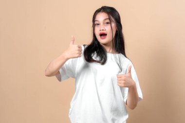 Heyecanlı genç Asyalı kadın beyaz bir tişört giyerek başparmağını kaldırırken ağzı açık bir şekilde gülümsüyor, stüdyo resminde bej arka planda duruyor..