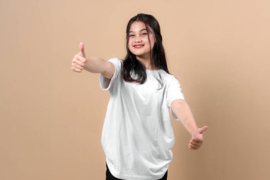 Beyaz tişörtlü neşeli genç Asyalı kadın, bej arka planda dikilirken güvenle gülümserken başparmağını kaldırıyor. Pozitif vücut dili ile stüdyo portresi.