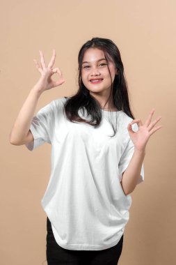 Beyaz tişört giyen, gülümseyen Asyalı genç bir kadının portresi ve iki eliyle bej bir arka plana karşı çift tamam el işareti yapıyor, pozitiflik ve güven gösteriyor..
