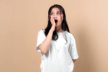Beyaz tişörtlü, tek eli ve ağzıyla yüzünü tutan Asyalı genç bir kadın, stüdyoda bej bir arka plana karşı şok ya da şaşkınlık ifadesi sergiliyor..