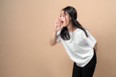 Beyaz tişörtlü ve siyah pantolonlu Asyalı bir kadın elleri ağızlarının yanında bağırıyor, bej rengi stüdyo arka planında yüksek sesle jest yaparken öne eğiliyor, anlamlı bir poz veriyor..