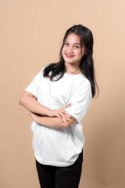 İki eliyle birden barış işareti yaparken parlak bir şekilde gülümseyen Asyalı genç bir kadının portresi. Beyaz bir tişört giyiyor ve bej bir arka plana karşı güvenle poz veriyor..