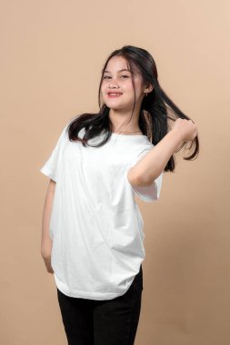 Uzun saçlarını tek eliyle tutarken gülümseyen kendinden emin genç Asyalı kadın. Sade beyaz tişört ve siyah pantolon giyiyor. Stüdyoda bej bir arka plana karşı duruyor..