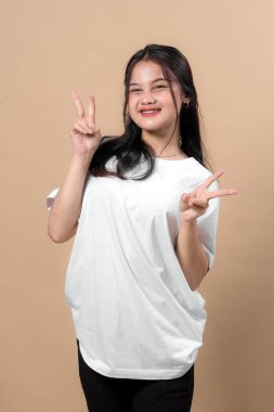 Düz beyaz tişört ve siyah pantolon giyen neşeli Asyalı genç bir kadın. Çifte barış işareti gösterirken parlak bir şekilde gülümsüyor. Bej renkli bir stüdyo arka planının önünde güvenle poz veriyor..