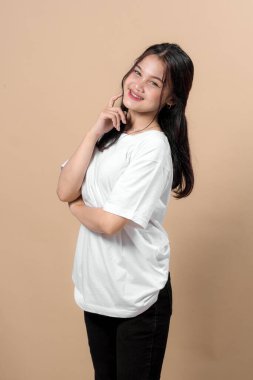 Uzun siyah saçlı çekici bir Asyalı kadın, sade beyaz tişört ve siyah pantolon giyiyor, bir eli çenesinde bej renkli bir arka planda poz verirken sıcak bir şekilde gülümsüyor..