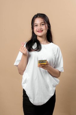 Genç Asyalı bir kadın kameraya gülümserken bir elinde akıllı bir telefon, diğerinde Koreli bir parmak kalp hareketi yapıyor, bej arka planda günlük kıyafetler giyiyor..