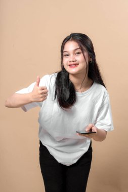 Asyalı genç bir kadın gülümserken baş parmak işareti yapıyor ve elinde akıllı bir telefon tutuyor. Hafifçe öne eğiliyor, bej arka planda günlük kıyafetler giyiyor..