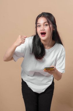 Neşeli, Asyalı genç bir kadın elinde akıllı bir telefonla gülümseyip yumruğunu kaldırıyor. Öne doğru eğiliyor, günlük beyaz tişört ve siyah pantolon giyiyor..