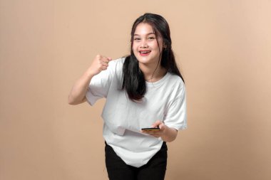 Neşeli, Asyalı genç bir kadın elinde akıllı bir telefonla gülümseyip yumruğunu kaldırıyor. Öne doğru eğiliyor, günlük beyaz tişört ve siyah pantolon giyiyor..