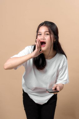 Genç Asyalı bir kadın elinde akıllı bir telefonla bağırıyor, beyaz tişört giyiyor, heyecanını dile getiriyor ya da bej arka planda bağırıyor..
