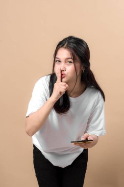 Genç Asyalı kadın sessiz bir jest yapıyor. Parmağını dudaklarına koyuyor, akıllı telefon tutuyor, beyaz tişört giyiyor, bej arka planda duruyor, ciddi görünüyor..