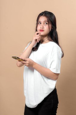 Sıradan beyaz tişörtlü düşünceli genç Asyalı kadın akıllı bir telefon tutuyor ve bej arka planda izole edilmiş çenesine dokunurken kafasını kaldırıyor. Fikir, ilham ya da karar kavramı.