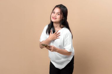 Beyaz tişörtlü, beyaz tişörtlü genç Asyalı bir kadın elinde akıllı bir telefonla hayret içinde, bej arka planda izole bir şekilde göğüslerine dokunuyor. Şok kavramı, vay be, ya da beklenmedik haberler..