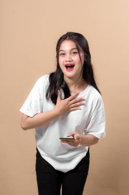 Beyaz tişörtlü, beyaz tişörtlü genç Asyalı bir kadın elinde akıllı bir telefonla hayret içinde, bej arka planda izole bir şekilde göğüslerine dokunuyor. Şok kavramı, vay be, ya da beklenmedik haberler..