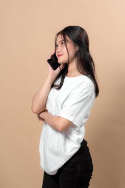 Akıllı bir telefonla konuşurken gülümseyen, kollarını kavuşturup poz veren kendinden emin, genç Asyalı bir kadın. İletişim ve bağlantı konsepti. Bej arka planda stüdyo çekimi.