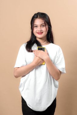 Genç Asyalı bir kadın, göğsüne yakın sarı bir akıllı telefon tutarken gülümsüyor, günlük beyaz bir tişört giyiyor, tarafsız bir bej arka plan karşısında duruyor..