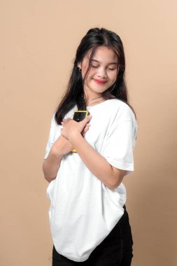 Kapalı gözlü genç Asyalı kadın göğsüne sarı bir akıllı telefonu sararken hafifçe gülümsüyor, beyaz bir tişört giyiyor, bej arka planda sakince duruyor..