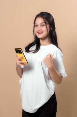 Genç Asyalı bir kadın, sarı bir akıllı telefon tutarken güvenle gülümsüyor, günlük beyaz tişört giyiyor, bej arka planda duruyor, neşeli görünüyor..