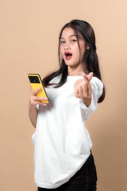 Asyalı genç bir kadının elinde sarı bir akıllı telefon tutması ve parmağıyla kalp hareketi yapması, günlük beyaz tişört giymesi, bej renkli stüdyo geçmişine karşı durması,.
