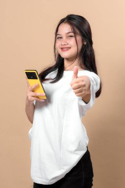 Beyaz tişörtlü gülümseyen Asyalı bir kadın elinde sarı bir akıllı telefon, baş parmak işareti yapıyor, onay ve pozitifliğini dile getiriyor, bej renkli stüdyo arka planına karşı duruyor..