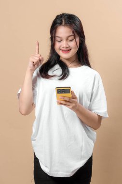 Beyaz tişörtlü gülümseyen Asyalı kadın elinde sarı akıllı telefon, işaret parmağıyla işaret ediyor, harika bir fikir ya da çözüm konsepti gösteriyor, bej arka planda duruyor..