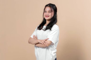 Kolları çapraz duran, kameraya gülümseyen kendinden emin genç Asyalı kadın. Beyaz tişörtlü, doğal makyajlı, bej arka planda izole bir kıyafet..