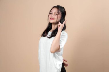 Neşeli genç Asyalı kadın işaret parmağıyla işaret ediyor, özgüvenle gülümsüyor. Beyaz bir tişört giyiyor ve stüdyoda bej bir arka plana karşı poz veriyor..